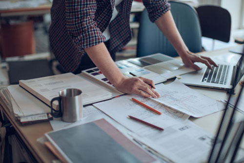 A person is standing over their desk working on paperwork and a laptop